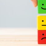 3 wooden blocks being stacked on top of each other: a red block with a frowning face, a yellow block with a neutral face, and a green block with a smiling face to represent peer review.