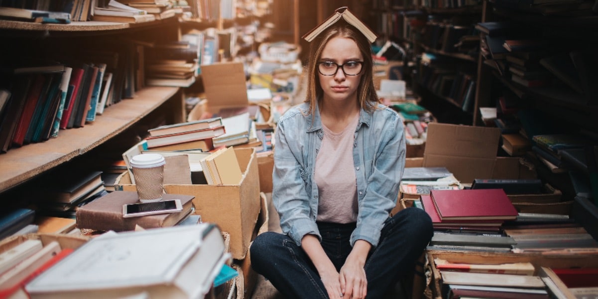 A young woman sitting on the floor of a library aisle surrounded by books and coffee and with an open book balanced on her head to represent keeping up with the literature
