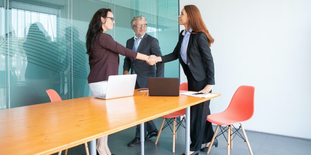 Two smiling women shaking hands and one smiling man standing in an interview room to represent getting a job after reading how to make a great impression at your life sciences interview