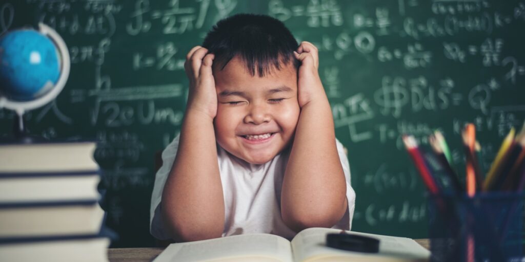 A young boy with hands on head surrounded by books and in front of a blackboard covered in scientific notations to represent preparing for the Qualifying Exam