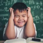 A young boy with hands on head surrounded by books and in front of a blackboard covered in scientific notations to represent preparing for the Qualifying Exam