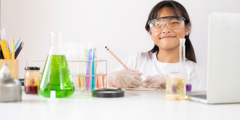 A photo of a young girl wearing lab coat and safety glasses surrounded by chemistry apparatus and writing in a book to represent organizing your lab book