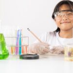 A photo of a young girl wearing lab coat and safety glasses surrounded by chemistry apparatus and writing in a book to represent organizing your lab book