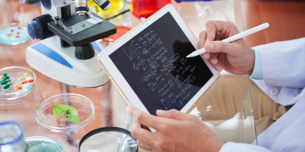 A picture of a scientist's hands holding a tablet with scientific notations surrounded by a microscope and scientific equipment to represent someone with a career in bioinformatics